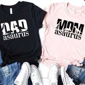 May include: Two black and pink t-shirts with a dinosaur graphic and the text "DAD asaurus" and "MOM asaurus". The shirts are laid out on a white wooden surface with two pairs of white sneakers and two pairs of blue jeans.