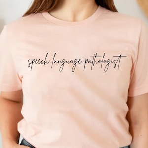 May include: A light pink t-shirt with the words "speech language pathologist" in a black script font. The shirt is a crew neck style and appears to be made of a soft material. The shirt is a solid color and has short sleeves.