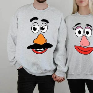 May include: A pair of grey sweatshirts with a cartoon face design. One sweatshirt features a Mr. Potato Head face with a black mustache and a brown nose. The other sweatshirt features a Mrs. Potato Head face with pink eyelashes and a pink nose.