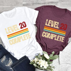 May include: Two t-shirts with a retro design. One is white with the text "Level 20 Complete" and the other is maroon with the text "Level 20 Complete".