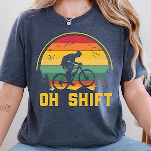 May include: Dark blue t-shirt featuring a cyclist silhouetted against a sunset design with the text "OH SHIFT" in yellow. The graphic includes a mountain bike, birds, and trees, creating a nature-themed design.