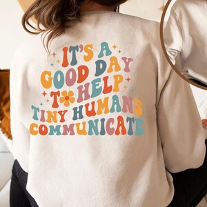 May include: A white sweatshirt with a retro-style graphic that says "It's a good day to help tiny humans communicate". The graphic is in rainbow colors. The sweatshirt also has a small graphic of an SLP logo on the front.