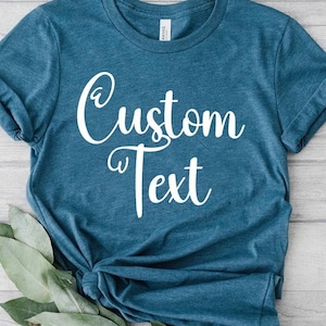 May include: A teal-colored t-shirt with the words "Custom Text" in white script. The shirt is knotted at the bottom and has rolled-up sleeves. Green leaves are placed at the bottom of the shirt.