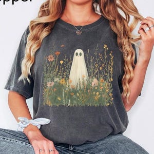 May include: Dark gray t-shirt featuring a whimsical ghost design amidst wildflowers. The ghost is a light cream color, and the flowers are in shades of yellow, orange, and green. The word "Pepper" is in the top left corner.