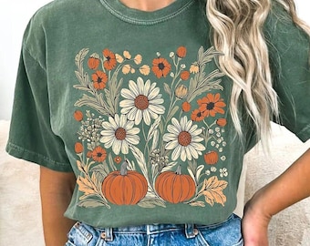 May include: A forest green short-sleeve t-shirt featuring a graphic print of two orange pumpkins surrounded by white daisies, orange flowers, and green foliage. The shirt has a crew neckline and a relaxed fit, showcasing a detailed botanical illustration.