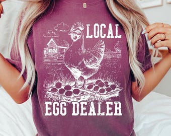 May include: A plum-colored t-shirt featuring a white graphic print of a chicken standing over two cartons of eggs in front of a barn. The text "LOCAL EGG DEALER" is printed in a bold, distressed white font above and below the central illustration.