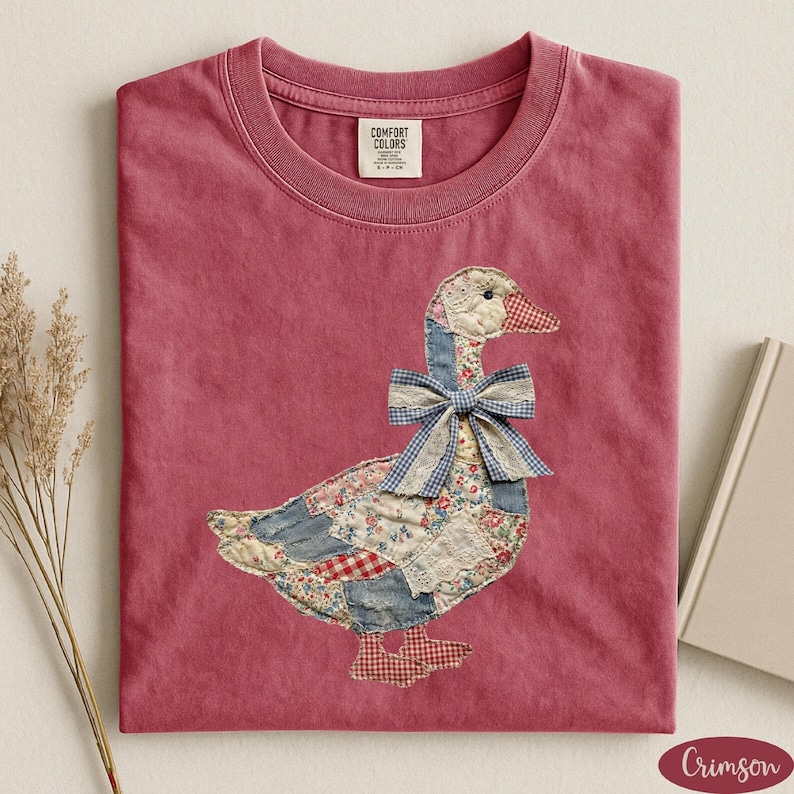 May include: A crimson t-shirt featuring a central patchwork goose graphic. The goose is constructed from various fabric patterns including blue denim, red gingham, and floral prints, and is adorned with a blue and white gingham bow at the neck.