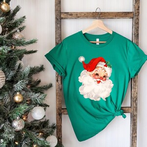 May include: A green t-shirt featuring a vintage Santa Claus graphic. The Santa Claus illustration has a red hat, white beard, and rosy cheeks. The shirt is hanging on a wooden ladder near a decorated Christmas tree.
