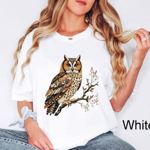 May include: White t-shirt featuring a detailed illustration of an owl perched on a branch. The owl has brown, tan, and white feathers, with large orange eyes. The branch has small leaves and the word "White" is printed on the bottom right.