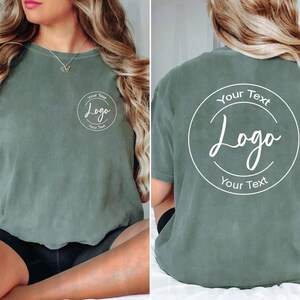 Puede incluir: Una camiseta de manga corta color verde salvia con un gr&aacute;fico circular blanco en el pecho izquierdo y una versi&oacute;n m&aacute;s grande del mismo gr&aacute;fico en la espalda. El gr&aacute;fico contiene el texto Your Text, Logo y Your Text en fuente blanca.