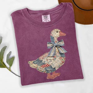 May include: A plum-colored t-shirt featuring a large goose graphic made of various patchwork fabric patterns including floral, denim, and red gingham. The goose wears a blue and white gingham bow around its neck. The shirt has a Comfort Colors label.