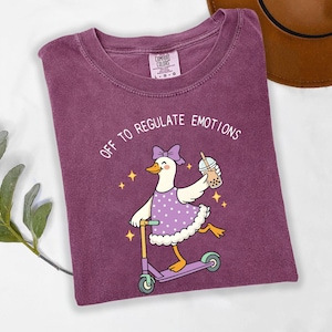 May include: A purple Comfort Colors t-shirt featuring a cartoon goose riding a scooter and holding a bubble tea. The goose wears a purple dress and bow, with the text "OFF TO REGULATE EMOTIONS" above the graphic.