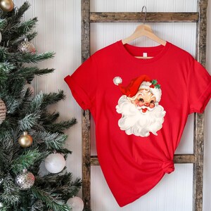May include: A red t-shirt featuring a vintage Santa Claus graphic. The Santa Claus illustration has a red hat, white beard, and a smiling face. The shirt is hanging on a wooden hanger.