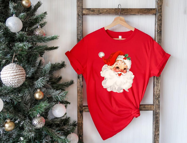 May include: A red t-shirt featuring a vintage Santa Claus graphic. The Santa Claus illustration has a red hat, white beard, and a smiling face. The shirt is hanging on a wooden hanger.