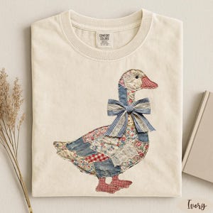 May include: An ivory t-shirt featuring a graphic of a goose made from a patchwork of floral, denim, and red-and-white checkered fabrics. The goose wears a blue-and-white gingham bow with lace trim around its neck. The shirt has a crew neckline.