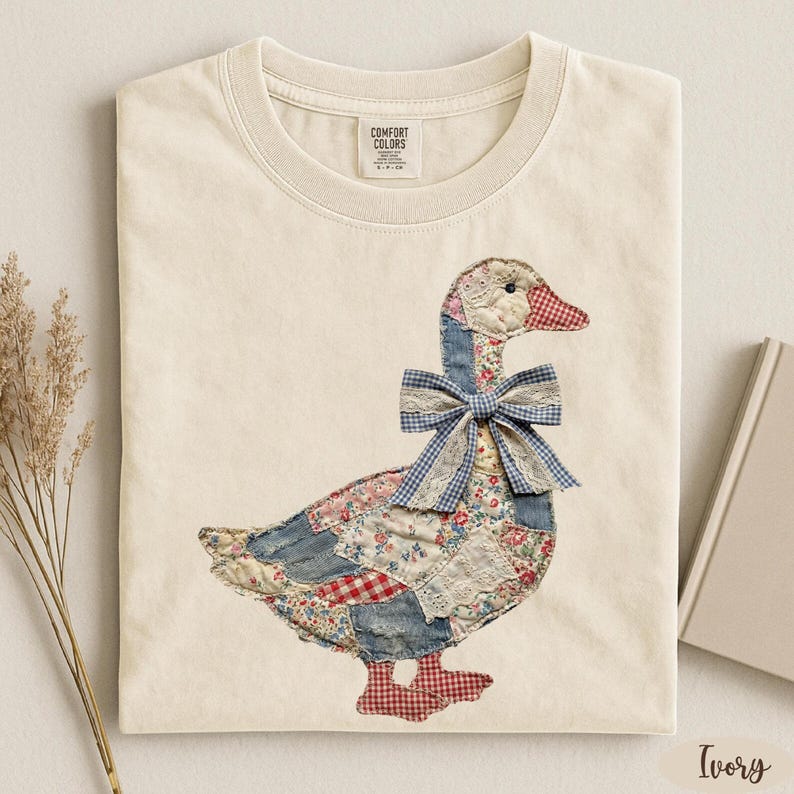 May include: An ivory t-shirt featuring a graphic of a goose made from a patchwork of floral, denim, and red-and-white checkered fabrics. The goose wears a blue-and-white gingham bow with lace trim around its neck. The shirt has a crew neckline.