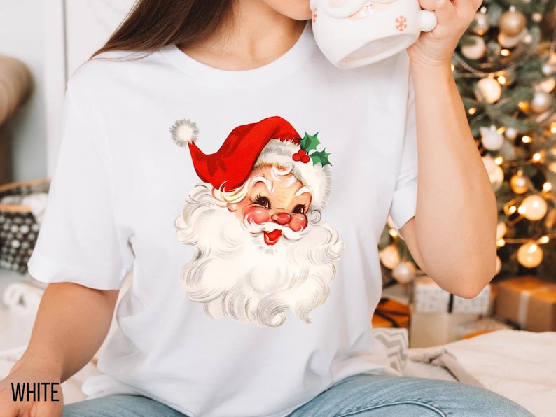 May include: White t-shirt featuring a vintage illustration of Santa Claus. Santa is depicted with a red hat, white beard, and rosy cheeks. The shirt has short sleeves and the word "WHITE" is printed at the bottom left.
