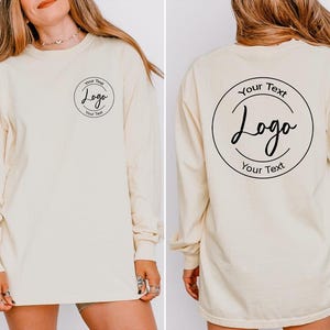 Puede incluir: Una camiseta de manga larga de color crema con un gr&aacute;fico circular negro en la parte frontal izquierda del pecho con el texto Your Text Logo Your Text. La parte posterior de la camiseta muestra una versi&oacute;n m&aacute;s grande del mismo gr&aacute;fico circular.