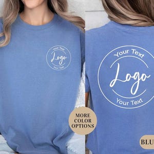 Puede incluir: Una camiseta azul de manga corta con un gr&aacute;fico circular blanco en el pecho izquierdo y una versi&oacute;n m&aacute;s grande del mismo gr&aacute;fico en la espalda. El gr&aacute;fico contiene el texto Your Text, Logo y Your Text dentro de un marco circular blanco.