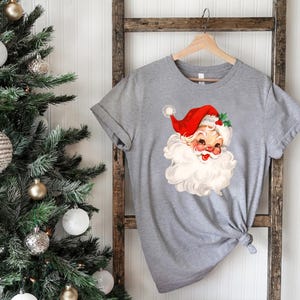 May include: A gray t-shirt featuring a vintage illustration of Santa Claus with a red hat, white beard, and rosy cheeks. The shirt is hanging on a wooden ladder near a decorated Christmas tree with white and gold ornaments. A festive holiday theme.