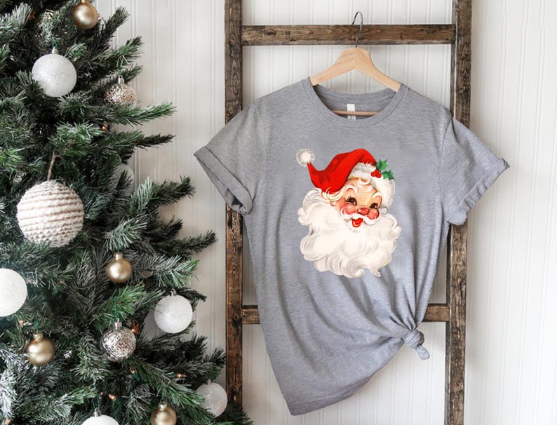 May include: A gray t-shirt featuring a vintage illustration of Santa Claus with a red hat, white beard, and rosy cheeks. The shirt is hanging on a wooden ladder near a decorated Christmas tree with white and gold ornaments. A festive holiday theme.