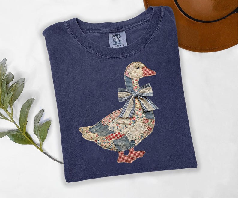 May include: A navy blue t-shirt featuring a large, central applique of a goose constructed from various fabric patches in floral, plaid, and solid patterns. The goose wears a blue and white gingham bow at its neck and has red plaid feet.