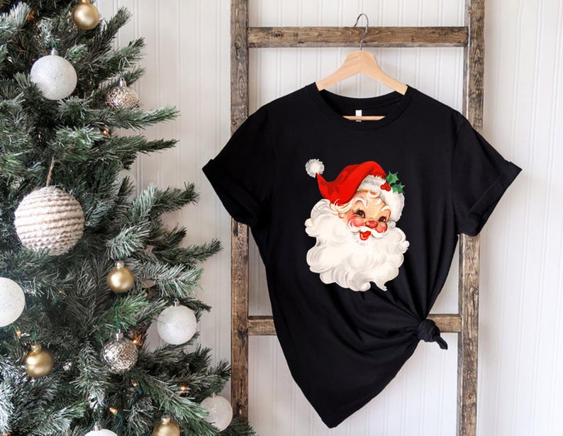 May include: Black t-shirt featuring a vintage Santa Claus graphic. The Santa illustration has a red hat, white beard, and rosy cheeks. The shirt is hanging on a wooden ladder near a decorated Christmas tree.
