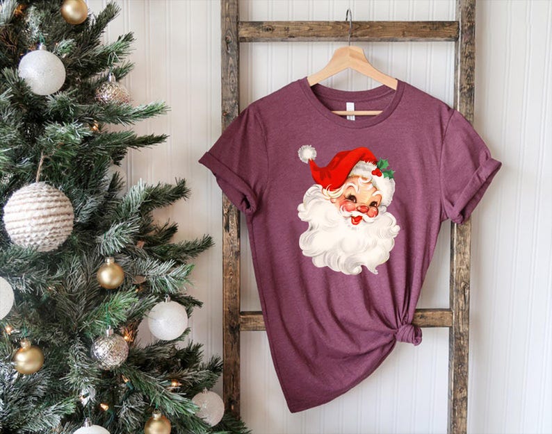 May include: A maroon t-shirt featuring a vintage Santa Claus graphic. The Santa illustration has a red hat, white beard, and rosy cheeks. The shirt is hanging on a wooden ladder near a decorated Christmas tree.