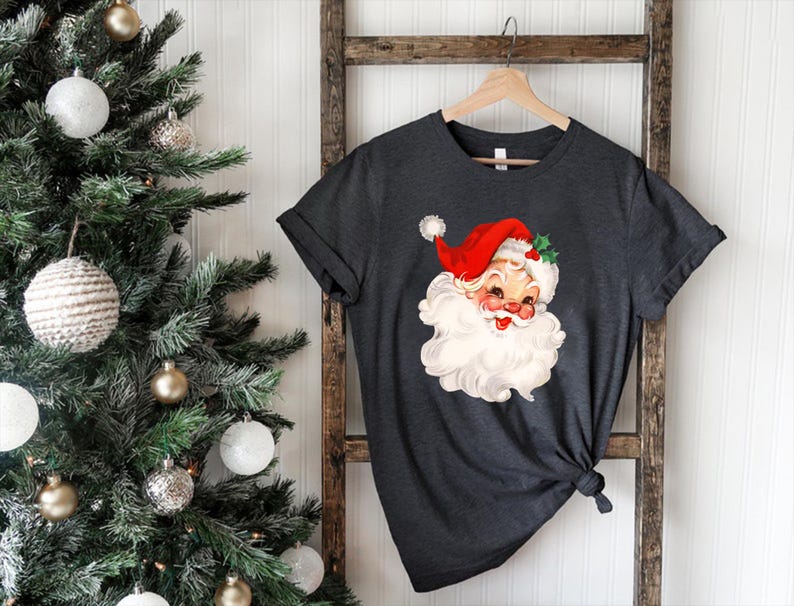 May include: Dark gray t-shirt featuring a vintage Santa Claus graphic. The Santa illustration has a red hat, white beard, and rosy cheeks. The shirt is hanging on a wooden ladder near a decorated Christmas tree.