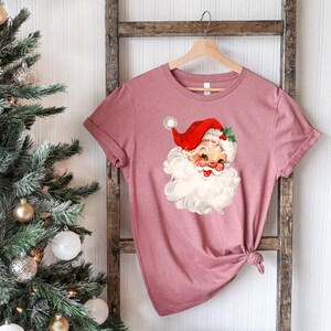 May include: A dusty rose-colored t-shirt featuring a vintage Santa Claus graphic. The Santa Claus illustration has a red hat, white beard, and rosy cheeks. The shirt is hanging on a wooden hanger against a white background.