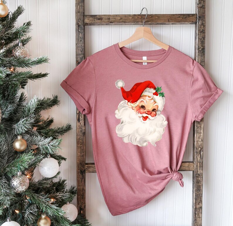 May include: A dusty rose-colored t-shirt featuring a vintage Santa Claus graphic. The Santa Claus illustration has a red hat, white beard, and rosy cheeks. The shirt is hanging on a wooden hanger against a white background.