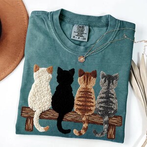 May include: Teal Comfort Colors t-shirt featuring four embroidered cats of different colors sitting on a wooden bench. The cats are white, black, brown, and gray. A gold necklace with a small pendant is draped on the shirt.