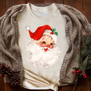 May include: Cream-colored t-shirt featuring a vintage Santa Claus graphic. Santa wears a red hat with white trim and holly accents. A brown cable-knit cardigan and pine sprigs with red berries and pinecones complete the festive scene.