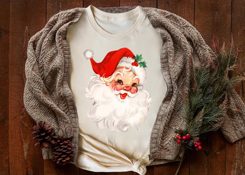May include: Cream-colored t-shirt featuring a vintage Santa Claus graphic. Santa wears a red hat with white trim and holly accents. A brown cable-knit cardigan and pine sprigs with red berries and pinecones complete the festive scene.