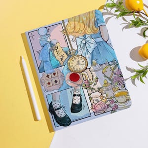 Può includere: Un quaderno con un'illustrazione ispirata ad Alice nel Paese delle Meraviglie. Il design include un orologio da tasca, tazze da tè e il testo "Drink Me". Uno stilo bianco è appoggiato su una superficie gialla.
