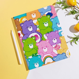 Peut inclure: Une coque de tablette colorée ornée de motifs d'ours de dessins animés dans diverses couleurs, notamment le violet, le vert, le rose et le bleu. Un stylet blanc est posé à côté de la coque sur une surface jaune. La coque présente un motif arc-en-ciel et cœur.