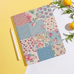 May include: A floral patchwork tablet case with a white stylus. The case features a variety of floral patterns in pink, blue, and cream colors. The case is rectangular and has a soft, fabric-like texture.