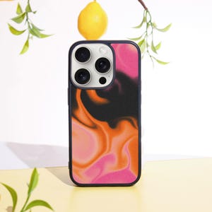 May include: A smartphone case with a black border and a vibrant abstract design in shades of pink, orange, and black. The case features a camera cutout with three lenses and a flash. A lemon and greenery are in the background.