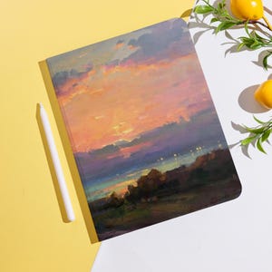 Könnte beinhalten: Ein Notizbuch mit einem Sonnenuntergangs-Landschaftsgemälde auf dem Einband. Das Kunstwerk zeigt lebendige Orange-, Rosa- und Lilatöne am Himmel, mit einer dunkelgrünen und braunen Landschaft darunter. Ein weißer Stift liegt neben dem Notizbuch.