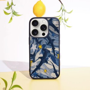 May include: A navy blue phone case featuring a pattern of hammerhead sharks, white stars, and yellow stars against a marbled blue and white background. The case has a black border and a camera cutout.