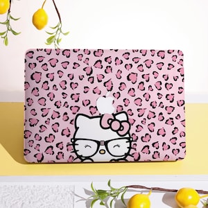 Könnte beinhalten: Rosa Laptop-Hülle mit einem Leopardenmuster aus schwarzen Herzen. Die Hülle zeigt einen weißen Cartoon-Katzencharakter mit Brille und Schleife. Das Apple-Logo ist oben sichtbar.
