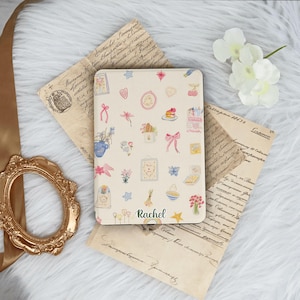 Könnte beinhalten: Eine cremefarbene Tablethülle mit skurrilen Illustrationen von Blumen, Schleifen und Lebensmitteln. Der Name "Rachel" ist in Schreibschrift geschrieben. Die Hülle liegt auf Vintage-Papier mit einem goldenen Rahmen und weißen Blumen.