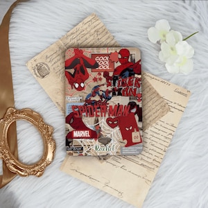 May include: A rectangular case with a Spider-Man design in red, blue, and white, incorporating comic book style elements and the Marvel logo. The name "Rachel" is also visible. The case is displayed on vintage paper and white fabric.