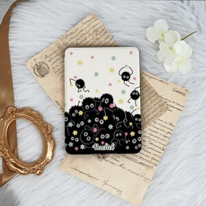 May include: A white tablet case featuring a whimsical design of black soot sprites with large eyes and colorful star accents. The name "Rachel" is written in cursive. The case is set against a backdrop of vintage ephemera.