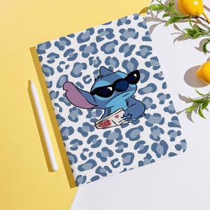 May include: A white tablet case with a blue leopard print pattern. The case features a cartoon illustration of Stitch wearing sunglasses and holding a card that says "LOVE LILO". A white stylus pen is on the left.