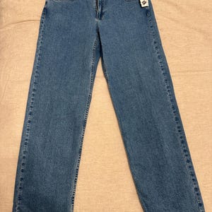 May include: Light blue denim jeans with a classic five-pocket design. The jeans have a straight leg cut and a zipper fly. A tag is attached to the front pocket. The jeans are laid flat on a neutral-colored surface.