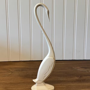 May include: A tall, off-white swan figurine featuring a long, curved neck and a stylized body with linear, embossed wing details. The swan stands on a small, hexagonal base, showcasing a smooth, solid-colored finish throughout the entire piece.