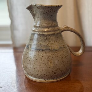 May include: A handmade ceramic pitcher with a speckled, earthy tone. The pitcher features a rounded body, a spout, and a curved handle. The neutral color and rustic design suggest a farmhouse or country aesthetic. The pitcher is likely used for serving beverages.