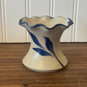 May include: A small, off-white ceramic vessel with a flared, ruffled rim. The piece features a hand-painted blue leaf motif on the exterior and a thin blue line accenting the scalloped edge of the opening. The surface has a speckled texture.