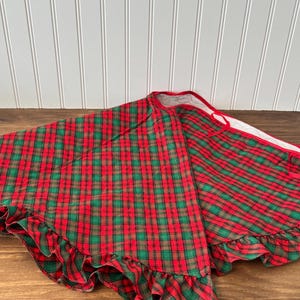 May include: A circular tree skirt featuring a red and green plaid pattern with a ruffled edge. The fabric has a red trim along the opening and a white lining visible on the underside. The design includes a slit for placement around a tree base.